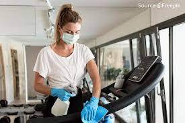 Person wearing gloves and a mask wiping down a treadmill using a spray bottle and blue rag. 