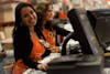 Woman with brown hair and olive skin smiling behind a computer register wearing an orange Home Depot apron.
