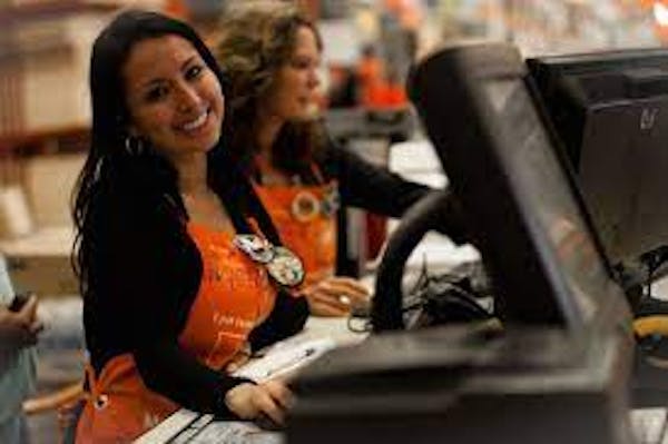 Woman with brown hair and olive skin smiling behind a computer register wearing an orange Home Depot apron.