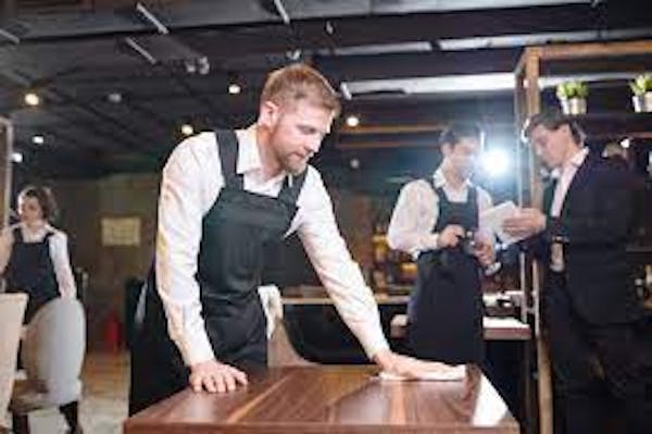 Person wearing a black apron wiping down a wood table with a rag with staff in the background.