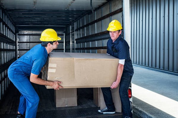 Two men wearing blue uniforms and yellow hard hats, lifting a box together