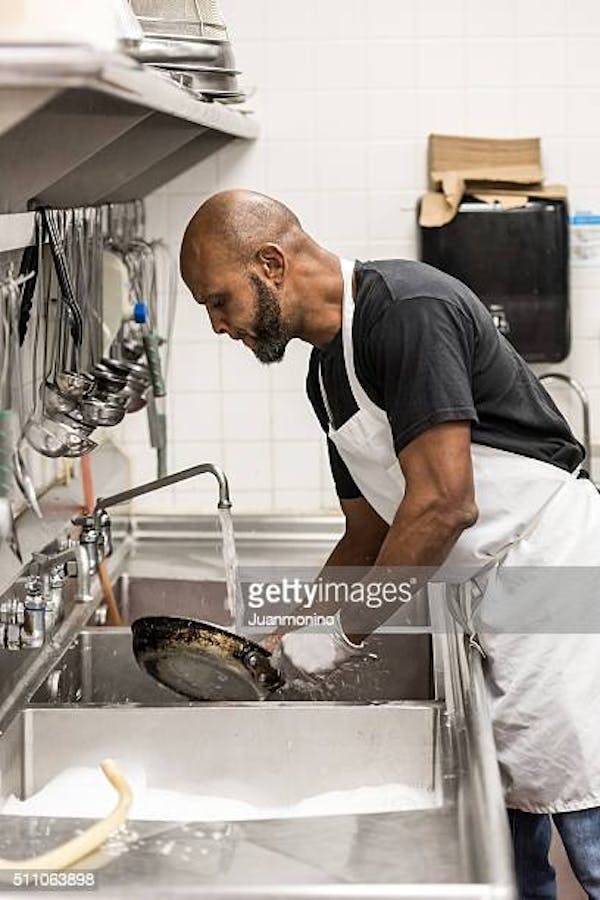 Man wearing a black shirt and white apron actively washing a cooking pan over a kitchen sink