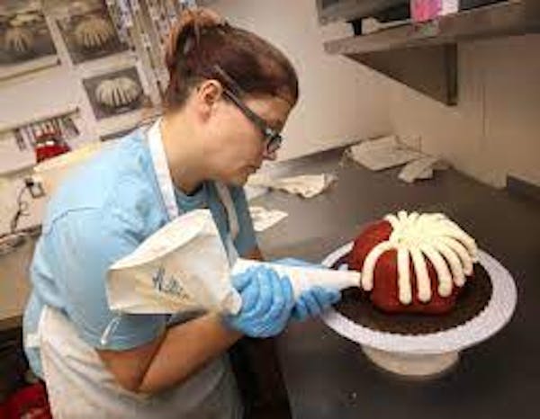Person wearing gloves frosting a red velvet bundt cake using a white piping bag and cake stand.