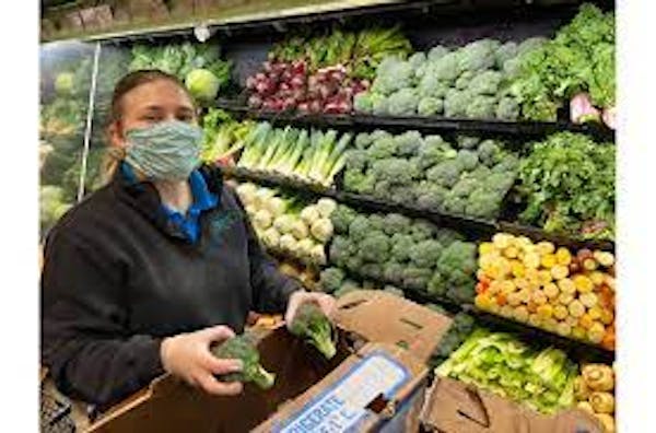 Person holding produce from a box with a mask on in front of the vegetable cooler.