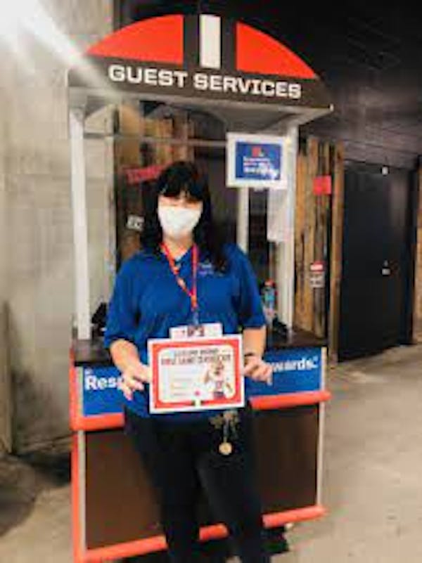 Person wearing face mask, blue shirt and black pants holding certificate in front of guest services kiosk.