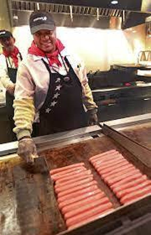 Person smiling while cooking multiple hotdogs on a flat griddle