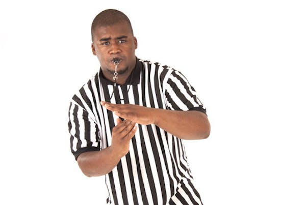 Man in a black & white striped referee shirt with a whistle in his mouth.