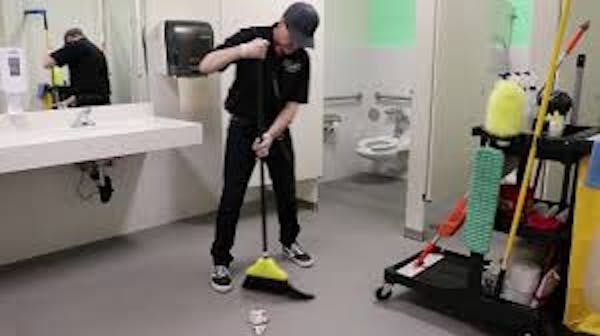 Person sweeping bathroom floors with a janitorial cart near by 