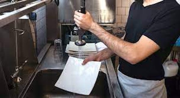 Person wearing a black t-shirt with a white apron, holding a white dish and using a sprayer to rinse the plate over a sink.