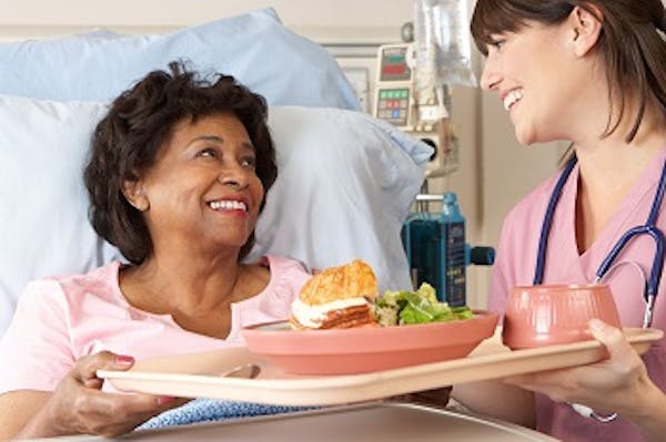 Person wearing scrubs delivering food tray to patient in hospital bed.