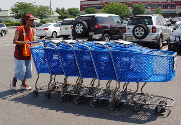 Person wearing an orange safety vest, pushing multiple blue shopping carts through a parking lot.