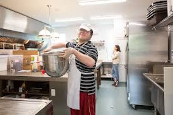 Person wearing a white apron and hat holding a large commercial sized mixing bowl in a bakery type environment,