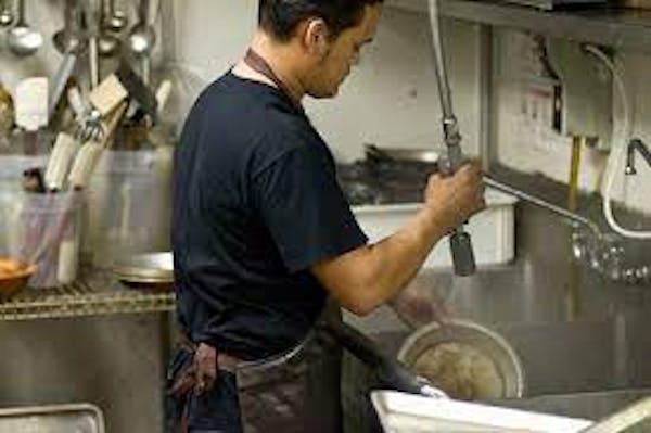Person holding a water hand-sprayer cleaning dirty plates in a large metal kitchen sink.