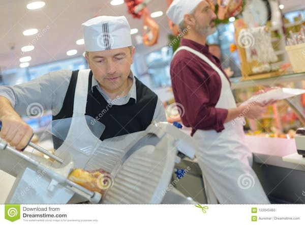 Person wearing a white, paper hat, wearing a blue button down shirt, with a white apron around his neck, covering his body.  The man is leaning over a metal, meat slicer, which holds a piece of deli meat,  In the background is another man wearing a burgundy shirt, wearing a white, paper hat and a white apron over his body.  He is holding a piece of meat on top of white parchment paper, while standing near a glass covered counter with foods inside.