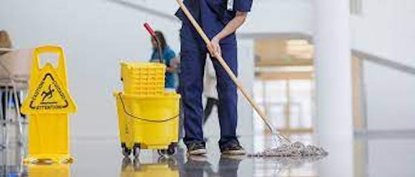 Person wearing a blue uniform while mopping a white floor, using a yellow mop bucket, and posting a yellow wet floor sign on the ground.