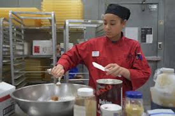 Person wearing a maroon shirt and black hat adding seasonings to a large mixing bowl