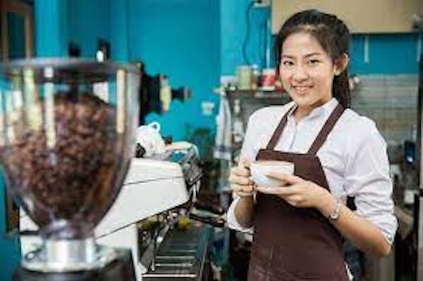 A person wearing a white shirt with a brown apron, holding a cup near a coffee station