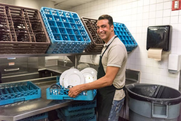 A white adult male with short brown hair wearing an untucked light grey polo t-shirt, blue jeans, a plain black apron. He is smiling, body slightly turned inward and resting a blue restaurant quality dish rack with assorted silverware and 3 white plates on a stainless steel surface. He's standing in a kitchen with white tiles and standing eye level with blue and brown dish racks. There's a black paper towel dispenser with a brown paper towel over a large trash bin with a black bag and grey base.