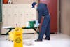 Male custodian mopping the floors in a school locker room with a yellow wet floor sign and yellow mop bucket on wheels. Cleaning supplies, paper towels, and a green bucket are sitting on the black bench in front of white lockers