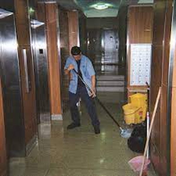 Person mopping a hallway in front of elevators with a yellow commercial mop bucket and extra mop leaning on the wall.
