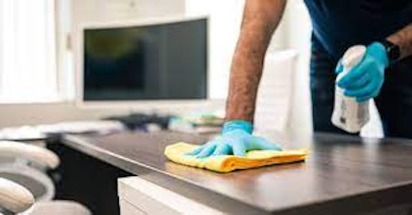 Picture of persons hands with a towel and spray bottle cleaning a desk with a computer on it.